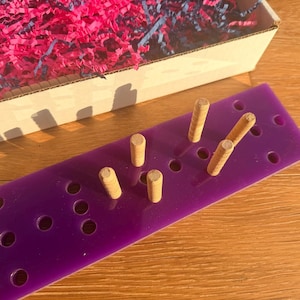 May include: A rectangular, purple pegboard with several wooden pegs inserted. The pegboard has evenly spaced holes. A cardboard box filled with pink and blue shredded paper sits in the background on a wooden surface.