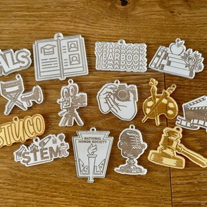 May include: A collection of wooden and acrylic charms with various school-related themes. Designs include a gavel, microphone, camera, and the words "PALS," "STUCO," "STEM," and "YEARBOOK." The charms are in shades of gold, silver, and white.