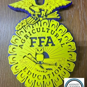 May include: A 3D-printed FFA emblem featuring a bright yellow eagle, owl, and plow design layered over a deep blue base. The circular emblem includes the text AGRICULTURAL EDUCATION and FFA in the center, with a small hanging loop at the top.