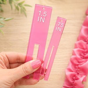 May include: Two pink acrylic ribbon gauges, one measuring 1.5 inches and the other 0.75 inches, are held in a hand. The gauges have a slot cut out of the center. A pink ribbon is in the background.