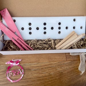 May include: A white box contains pink tools, wooden dowels, and a white board with black holes. The box sits on a wooden surface, with a pink and white FFA emblem and a silver medal with a blue ribbon.