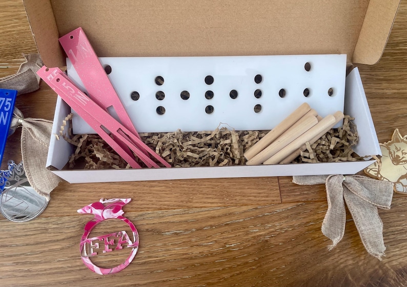 May include: A white box contains pink tools, wooden dowels, and a white board with black holes. The box sits on a wooden surface, with a pink and white FFA emblem and a silver medal with a blue ribbon.