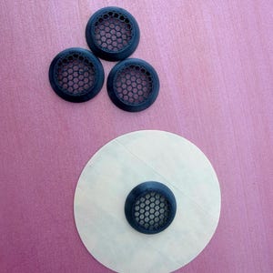 May include: Three dark blue, circular vent covers with a honeycomb pattern are arranged on a pink surface. One vent cover is attached to a white, circular sticker. The vent covers are made of plastic.