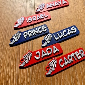 May include: A collection of personalized name tags in red and blue, each with a white name and a silver winged emblem. Names include Anaya, Israel, Prince, Lucas, Jada, and Carter. The tags are arranged on a wooden surface.