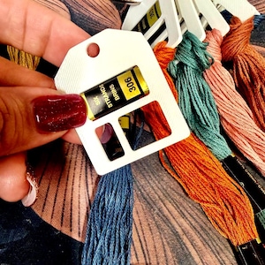 May include: A collection of embroidery floss skeins in various colors including blue, orange, teal, pink, and brown, each attached to a white plastic organizer tag labeled with the text Six Strands Brins and specific color numbers.