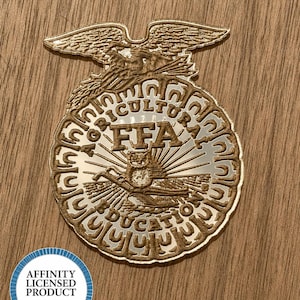 May include: A metallic gold-toned FFA emblem featuring an eagle at the top, a central owl, and a plow, with the text AGRICULTURAL EDUCATION and FFA embossed on the surface. A circular blue and white sticker reads AFFINITY LICENSED PRODUCT.