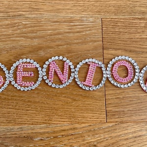 May include: Pink rhinestone letters spell out the word "SENIOR" on a wooden surface. Each letter is outlined with clear rhinestones. The letters are in a sans-serif font and are evenly spaced.