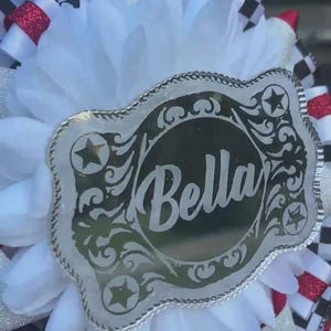 May include: A decorative item featuring a silver-colored buckle with the name "Bella" in a script font. The buckle is surrounded by white fabric, red glitter accents, and black and white checkered ribbons. The item has a festive appearance.