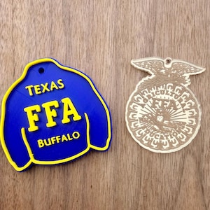 May include: A blue jacket-shaped ornament with yellow trim and the text TEXAS FFA BUFFALO, alongside a clear acrylic circular ornament featuring an eagle, an owl, and the text AGRICULTURAL EDUCATION FFA, both with small holes for hanging.