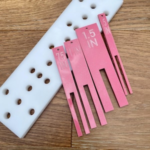 May include: A white foam block with evenly spaced holes and several pink, glittery, slotted tools. The tools are labeled with measurements: 0.5 IN, 0.75 IN, and 1.5 IN. The tools are used for crafting or DIY projects.