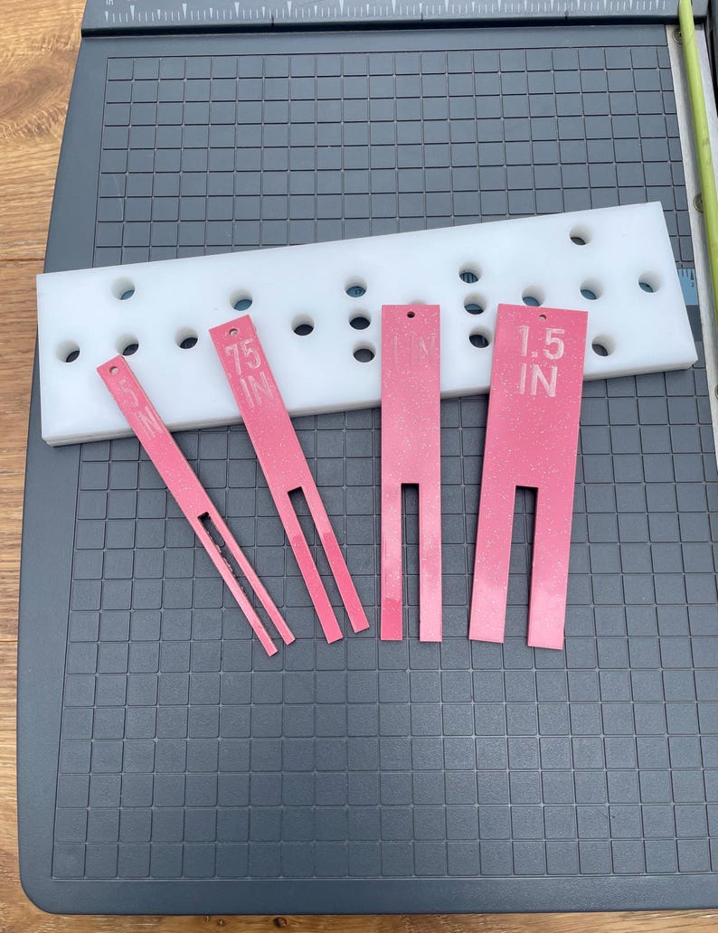 May include: A white plastic guide with multiple holes and four pink tools. The tools are labeled with measurements of 1.5 IN and 0.75 IN. The tools have a slotted design.