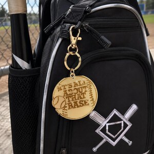 May include: A gold-colored baseball-themed keychain with the phrase "IT'S ALL ABOUT THAT BASE" engraved on it. The keychain is attached to a black backpack with a baseball bat in the side pocket. The backpack has a baseball diamond and crossed bats design.
