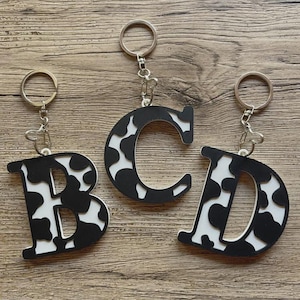 May include: Three letter-shaped keychains featuring the letters B, C, and D. Each keychain is made of a white material with a black cow-print pattern on the front and includes a silver-toned metal split ring attached by a small chain link.