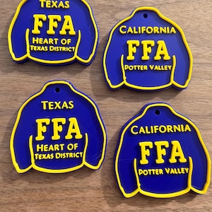 May include: Four blue and yellow jacket-shaped ornaments with a hole at the top. Two ornaments feature the text 'TEXAS FFA HEART OF TEXAS DISTRICT' and two feature the text 'CALIFORNIA FFA POTTER VALLEY' in raised yellow lettering on a blue background.