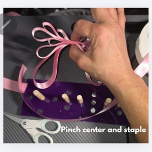 May include: A pink ribbon bow being crafted with a purple bow maker. The bow maker has wooden pegs and a curved design. Scissors are visible in the foreground. The text "Pinch center and staple" is at the bottom.