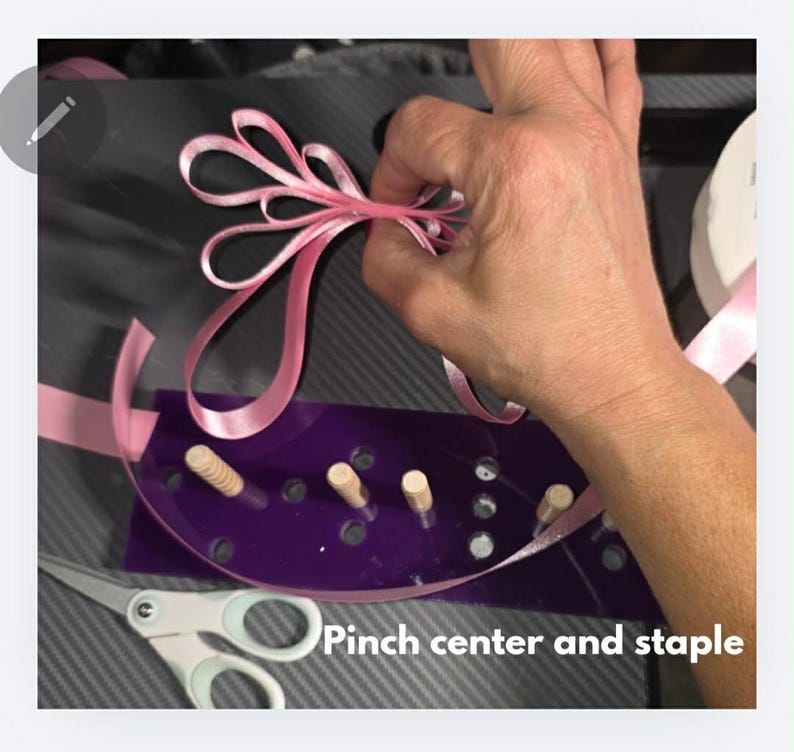 May include: A pink ribbon bow being crafted with a purple bow maker. The bow maker has wooden pegs and a curved design. Scissors are visible in the foreground. The text "Pinch center and staple" is at the bottom.