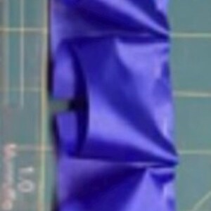 May include: Close-up of a vibrant blue ribbon with a unique, folded design. The ribbon is arranged in a repeating pattern, creating a textured effect. The background is a green cutting mat with measurement markings.