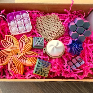 May include: A gift box filled with various colorful fidget toys and accessories. Includes a purple and white pop-it, a flower-shaped ornament, and a geometric cube. The box is lined with bright pink shredded paper.