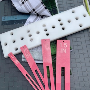 May include: A white plastic quilting template with multiple holes, pink measuring tools labeled .5 IN, .75 IN, and 1.5 IN, and fabric samples with plaid and green patterns. The tools are for measuring and cutting fabric.