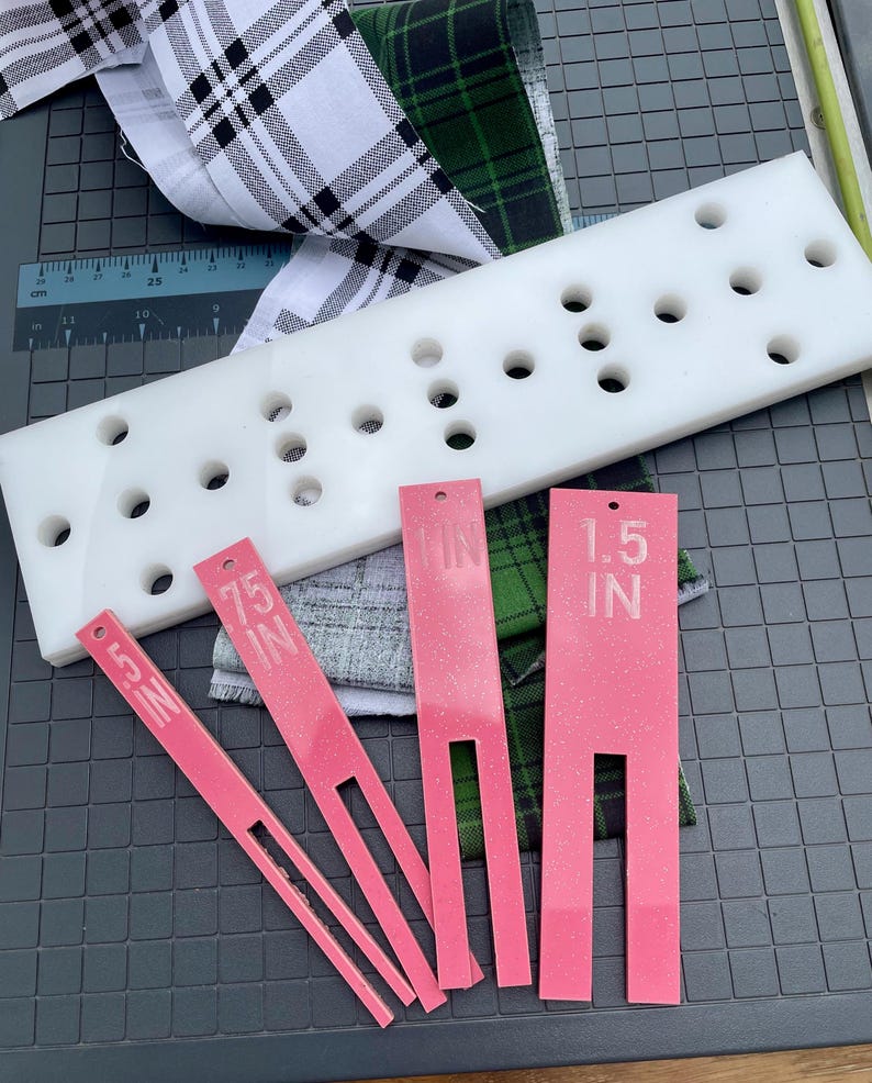 May include: A white plastic quilting template with multiple holes, pink measuring tools labeled .5 IN, .75 IN, and 1.5 IN, and fabric samples with plaid and green patterns. The tools are for measuring and cutting fabric.