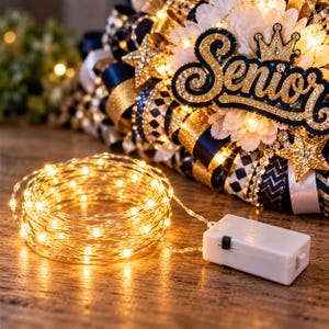 May include: A coil of warm white LED fairy lights on a thin silver wire with a white rectangular battery pack featuring a black power switch, positioned in front of a decorative wreath with navy blue, gold, and white ribbons and a Senior sign.