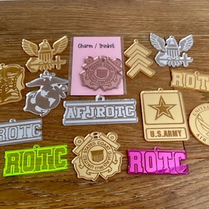 May include: A collection of military-themed charms and trinkets in various shapes and colors. Includes gold, silver, and neon green acrylic pieces with text like "ROTC", "U.S. Army", and "Air Force". A pink card reads "Charm / Trinket".