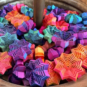 May include: A collection of colorful, star-shaped objects in shades of pink, orange, blue, green, and purple. The objects are layered, creating a three-dimensional effect. They are contained within a metal container.