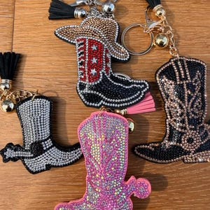 May include: Four boot-shaped keychains decorated with rhinestones. One features a red and black boot with a hat, one is black with gold accents, one is silver, and one is pink. Each includes a gold-tone metal keyring and a small tassel.