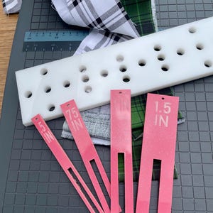 May include: A white plastic quilting ruler with multiple holes, alongside pink measuring tools labeled "1.5 IN" and ".75 IN". Plaid fabric in black and white, and green and black are also present. The items are on a gray cutting mat.