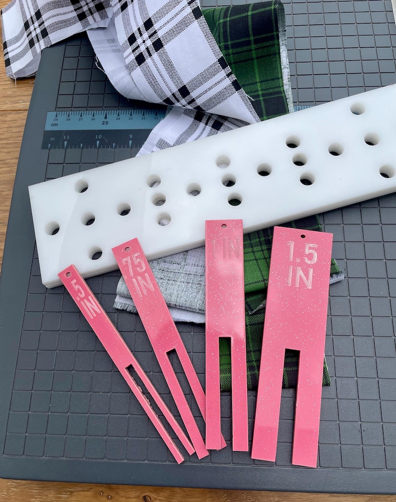 May include: A white plastic quilting ruler with multiple holes, alongside pink measuring tools labeled "1.5 IN" and ".75 IN". Plaid fabric in black and white, and green and black are also present. The items are on a gray cutting mat.