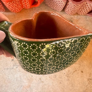 May include: A green heart-shaped ceramic mug with white polka dots. The mug has a handle and is made of a rustic, hand-crafted style.