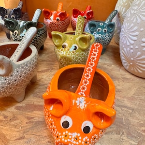 May include: A selection of ceramic pig-shaped bowls with spoons. The bowls are painted in various colours, including orange, green, and grey, and decorated with white polka dots. The spoons have a floral design.