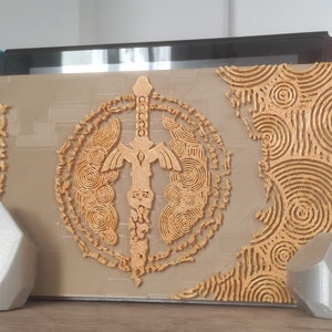 Peut inclure: Un support d'impression 3D pour une console Nintendo Switch, avec un design doré orné d'une épée et de motifs circulaires. Le support est en matériau gris clair et a une forme géométrique.
