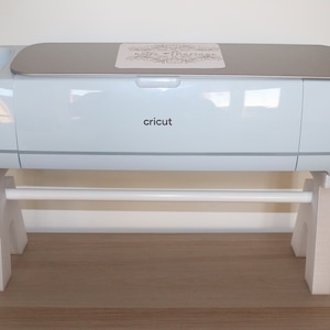 Può includere: Una macchina da taglio Cricut Explore Air 2 bianca con un top argentato e un supporto per rotoli bianco. La macchina è su una superficie di legno.