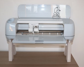 Könnte beinhalten: Eine hellgraue Cricut Maker 3 Maschine mit geöffnetem Deckel, die die Schneideklinge und die Matte zeigt. Die Maschine steht auf einem hellen Holzständer. Auf dem Deckel befindet sich ein Cartoon-Käfer-Aufkleber.