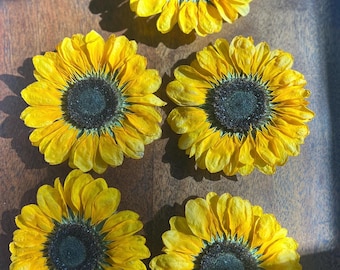 Può includere: Cinque girasoli giallo brillante con centri scuri sono disposti su una superficie di legno. I petali sono completamente aperti, mostrando il loro colore vibrante. I girasoli sono il punto focale, creando un'estetica allegra e naturale.
