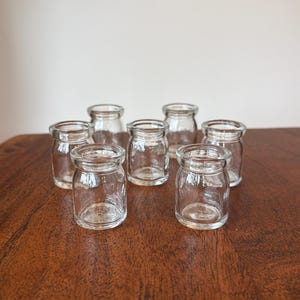 May include: A collection of seven small, clear glass jars with a rounded body and a slightly flared rim. Each jar features a smooth, transparent surface and a wide opening, arranged in a cluster to showcase their uniform shape and design.