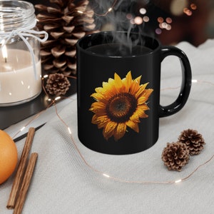 May include: Black coffee mug featuring a yellow sunflower design. The sunflower has water droplets on its petals.