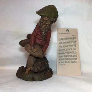 May include: A whimsical figurine of a gnome sitting on a mushroom with a small hedgehog at his feet. The gnome is wearing a red shirt, green hat, and has a long white beard. The figurine is made of a brown, textured material. The text on the card reads "Kevin and Cora #51:52".