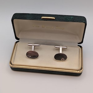 May include: Silver cufflinks with dark grey oval faces, each with a starburst design. The cufflinks are displayed in a green, textured box. The box interior is cream-colored, and the text "The Watch Shop" is visible.