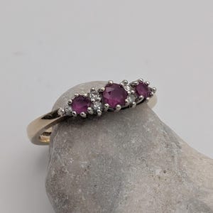 May include: A gold ring with a band featuring five red gemstones and small, clear stones. The ring is displayed on a light-coloured stone, highlighting the jewellery's design and colour contrast. The ring's band is a simple, polished gold.