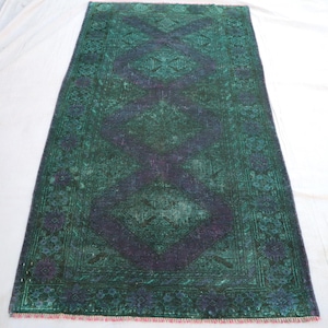 Puede incluir: Una alfombra rectangular, desgastada, en tonos verdes y morados. La alfombra presenta un patrón central de diamantes y un borde floral. La alfombra tiene un aspecto descolorido y un borde con flecos. Probablemente sea de lana o material similar.