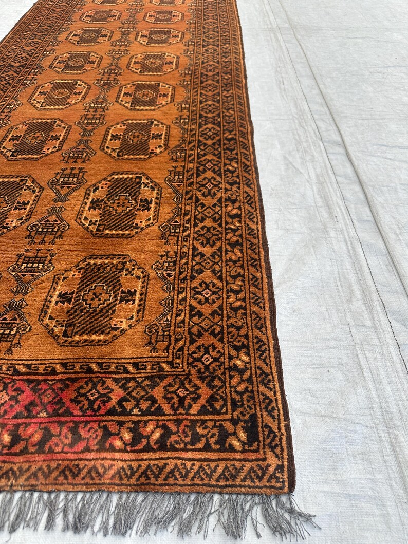 May include: A rectangular runner rug featuring a warm orange background with repeating dark brown octagonal geometric motifs. The border displays intricate black and orange patterns, and the short end is finished with grey fringe tassels.