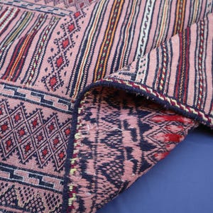 May include: A woven textile featuring a dusty pink base with intricate geometric patterns in dark blue, red, and white. The fabric displays a combination of vertical stripes and diamond motifs, with a folded corner revealing the reverse side.