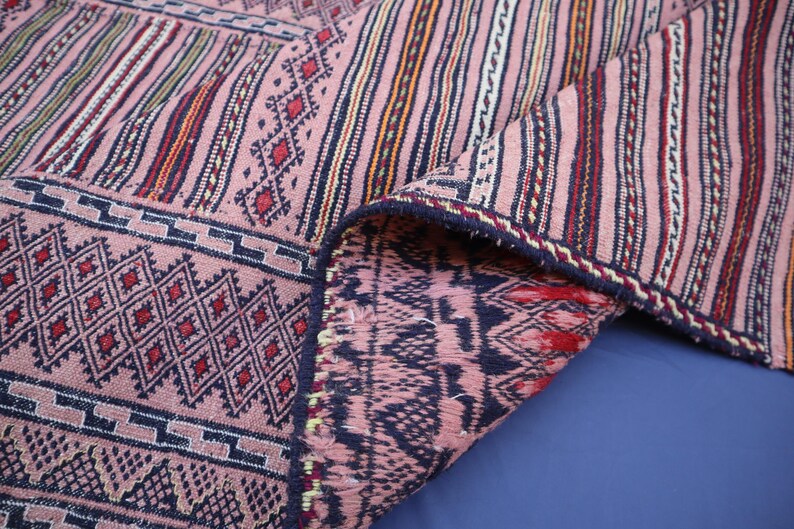 May include: A woven textile featuring a dusty pink base with intricate geometric patterns in dark blue, red, and white. The fabric displays a combination of vertical stripes and diamond motifs, with a folded corner revealing the reverse side.
