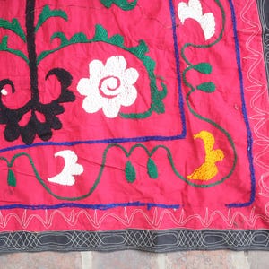K&ouml;nnte beinhalten: Nahaufnahme eines leuchtend roten Textils mit aufwendiger Stickerei. Das Design zeigt wei&szlig;e Blumenmuster, gr&uuml;ne Bl&auml;tter und ein schwarzes zentrales Motiv. Ein Rand mit einem grau-wei&szlig;en geometrischen Muster umrahmt die Kanten.