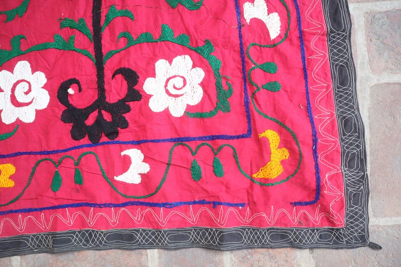 K&ouml;nnte beinhalten: Nahaufnahme eines leuchtend roten Textils mit aufwendiger Stickerei. Das Design zeigt wei&szlig;e Blumenmuster, gr&uuml;ne Bl&auml;tter und ein schwarzes zentrales Motiv. Ein Rand mit einem grau-wei&szlig;en geometrischen Muster umrahmt die Kanten.