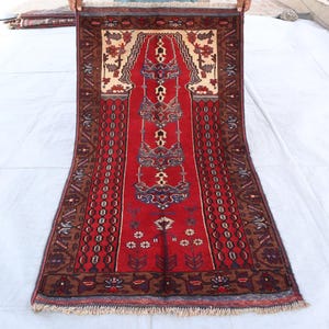 May include: A red and brown prayer rug with intricate floral and geometric patterns. The rug has a fringe border and is made of wool.