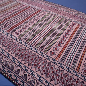 May include: A rectangular woven textile runner featuring a soft pink base color. The design consists of repeating geometric patterns, including dark blue diamond shapes, intricate zig-zag borders, and vertical stripes in red, white, and black.