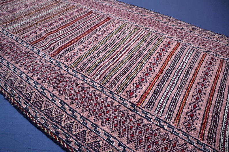 May include: A rectangular woven textile runner featuring a soft pink base color. The design consists of repeating geometric patterns, including dark blue diamond shapes, intricate zig-zag borders, and vertical stripes in red, white, and black.